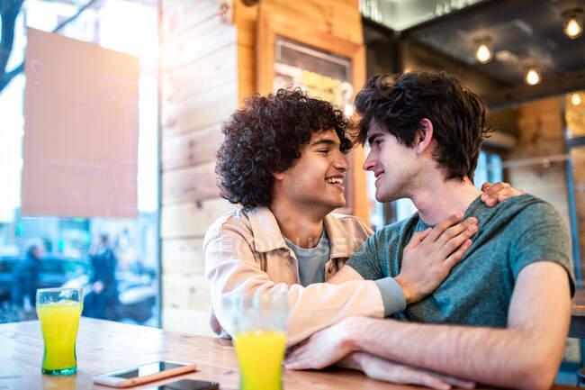 Vue latérale d'hommes ethniques excités s'embrassant sur la table et riant pendant un rendez-vous romantique dans la cafétéria moderne — Photo de stock