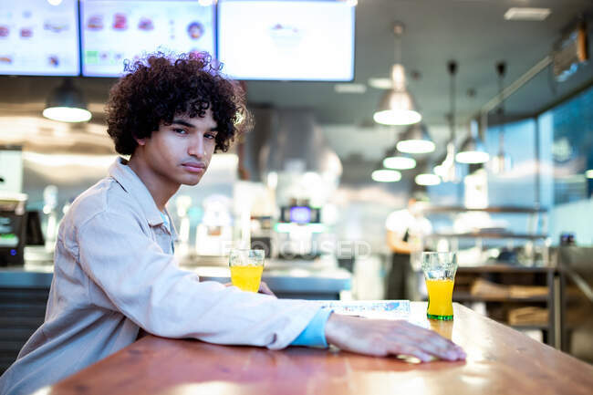 Vista lateral del joven con el pelo rizado mirando a la cámara y esperando la fecha mientras está sentado en la mesa con vasos de jugo en la cafetería - foto de stock