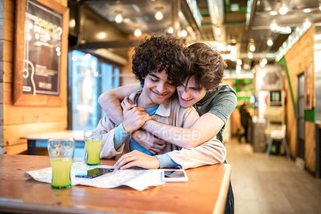 Hombres homosexuales jóvenes multiétnicos abrazándose mirando hacia el mapa de navegación y tomando bebidas frescas sonriendo mientras están sentados en la mesa de la cafetería durante una cita romántica - foto de stock
