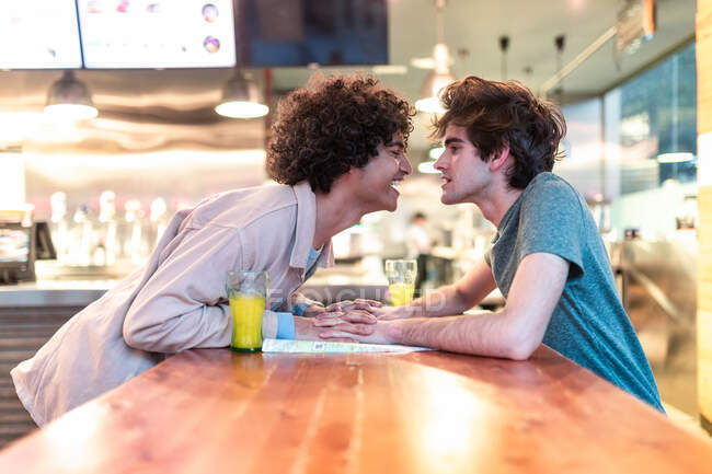 Side view of excited ethnic men embracing each other holding hands over table and laughing during romantic date in modern cafeteria — Stock Photo