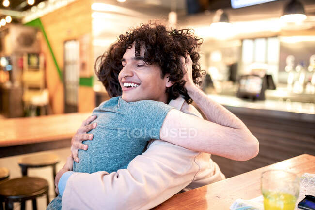 Excited ethnic men embracing each other with closed eyes on table and laughing during romantic date in modern cafeteria — Stock Photo