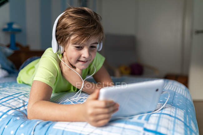 Niño preadolescente positivo en ropa casual con auriculares acostados en la cama y viendo películas en la tableta mientras descansa en casa - foto de stock