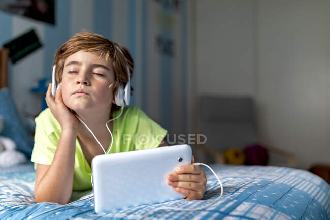 Menino pré-adolescente positivo em desgaste casual com fones de ouvido deitados na cama e assistindo filme em tablet enquanto descansa em casa — Fotografia de Stock