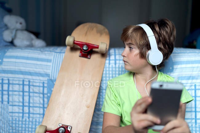 Niño pequeño con auriculares escuchando música y charlando con amigos en la red social mientras está sentado cerca de monopatín en el dormitorio - foto de stock