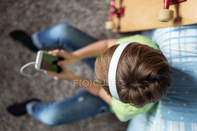 Petit garçon avec écouteurs écouter de la musique et bavarder avec des amis dans le réseau social tout en étant assis près de planche à roulettes dans la chambre — Photo de stock