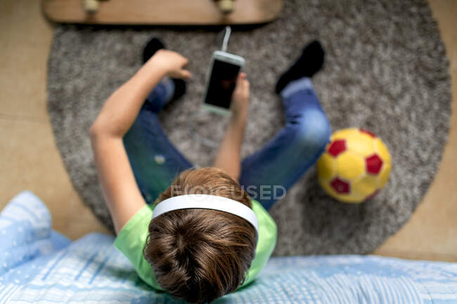Little boy with headphones listening to music and chatting with friends in social network while sitting near skateboard in bedroom — Stock Photo