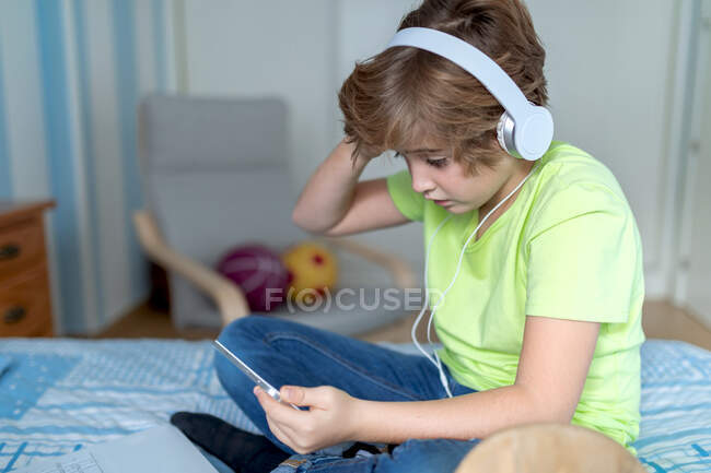 Colegial serio en traje casual y auriculares jugando videojuego en la tableta en casa - foto de stock