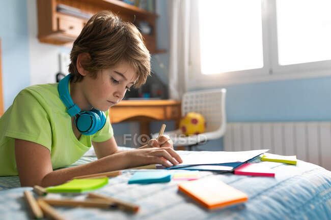 Продуманий школяр у повсякденному носінні та бездротових навушниках насолоджується музикою та малюнком з олівцями, проводячи вільний час у спальні — стокове фото
