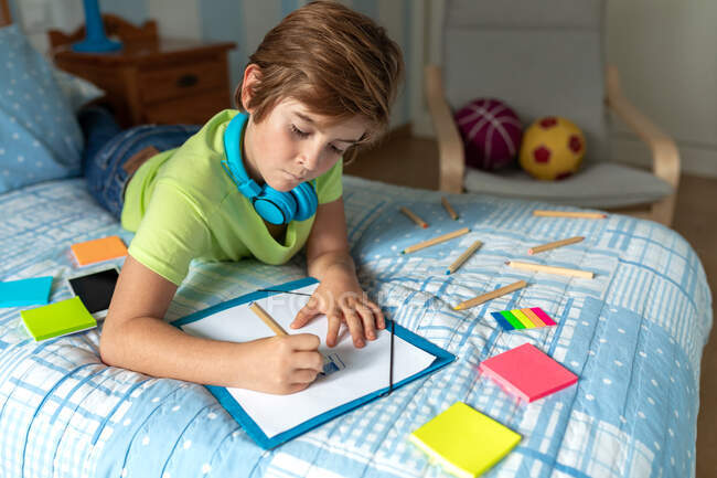 Thoughtful schoolboy in casual wear and wireless headphones enjoying music and drawing with pencils while spending free time in bedroom — Stock Photo
