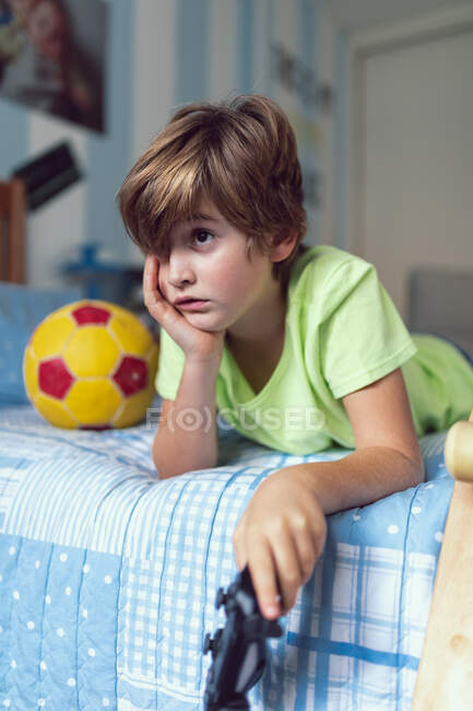 Bored little boy in casual clothes lying on bed near ball and skateboard unhappy with self isolation at home — Stock Photo