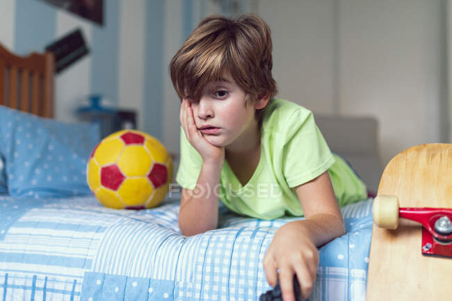 Bored  little boy in casual clothes lying on bed near ball and skateboard unhappy with self isolation at home — Stock Photo