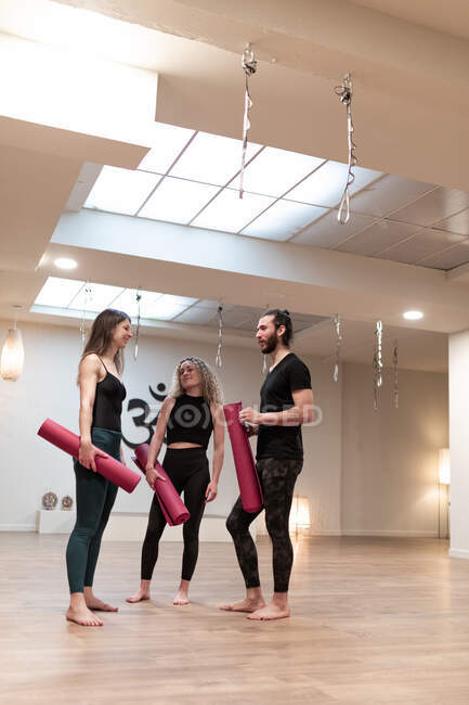 Молодий чоловік і жінки з килимками розмовляють один з одним, стоячи в просторій студії під час тренування з йоги — стокове фото