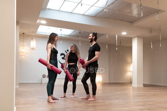 Jovem e mulheres com esteiras falando uns com os outros enquanto estão em estúdio espaçoso durante o treinamento de ioga — Fotografia de Stock