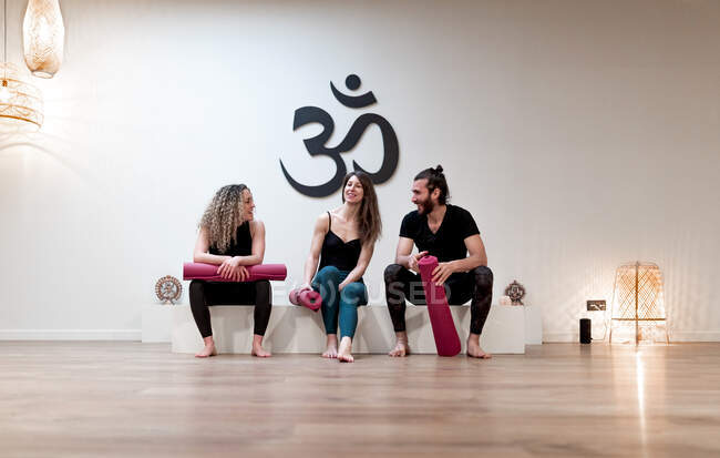Jeune homme et femmes avec des nattes parlant entre eux tout en étant assis dans un bloc blanc dans un studio spacieux pendant la formation de yoga — Photo de stock