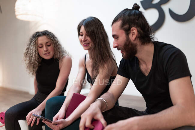 Joyeux jeune femme souriant et montrant les données sur smartphone aux amis enchantés avec des nattes avant l'entraînement de yoga en studio — Photo de stock