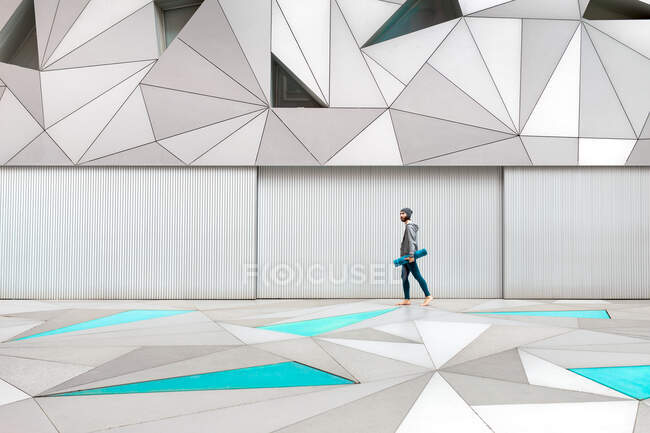 Ragazzo barbuto cercando modo mentre a piedi e tenendo tappetino yoga sotto il braccio vicino parete metallica in edificio moderno con soffitto ottagonale — Foto stock