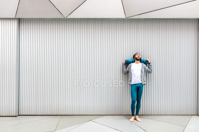 Bearded guy with closed eyes stretching and massaging neck with rolled mat while leaning on wall during yoga training — Stock Photo