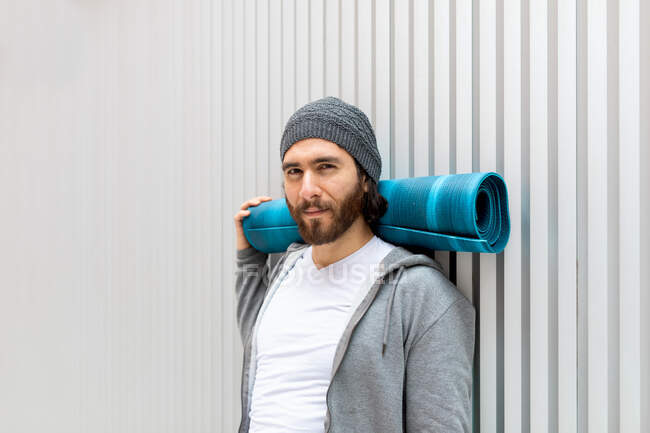 Bearded guy looking at camera with rolled mat supporting neck while leaning on wall during yoga training — Stock Photo