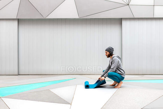 Ragazzo barbuto accovacciato guardando altrove mentre srotolava tappetino yoga sul pavimento dopo la lezione di yoga vicino alla parete metallica in un edificio moderno con soffitto ottagonale — Foto stock