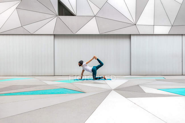 Vue latérale d'un homme adulte en tenue de sport faisant de l'exercice de pose de tigre tout en faisant du yoga contre un mur géométrique dans une pièce spacieuse — Photo de stock
