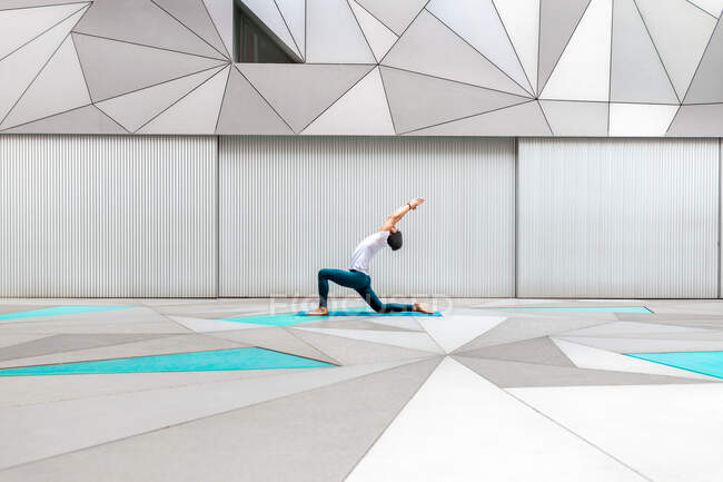 Vista laterale di maschio adulto in abbigliamento sportivo facendo basso affondo posa esercizio yoga in camera spaziosa con parete geometrica e pavimento — Foto stock