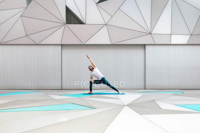 Ganzkörpererwachsener Mann in Sportbekleidung, der in einem geräumigen Raum mit geometrischer Wand und Boden seitliche Winkelübungen macht — Stockfoto