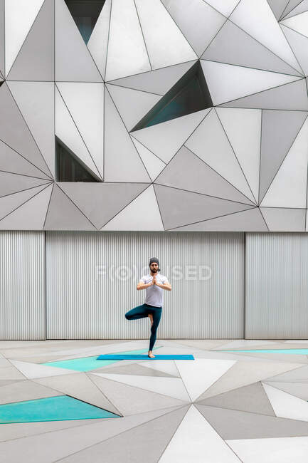 Hombre adulto de cuerpo completo en ropa deportiva realizando ejercicio de pose de árbol de pie mientras hace yoga contra la pared con adorno geométrico - foto de stock