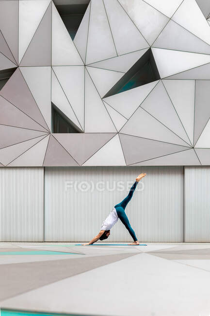 Hombre adulto de cuerpo completo en ropa deportiva realizando ejercicio Three Legged Downward Facing Dog mientras hace yoga contra la pared con adorno geométrico - foto de stock