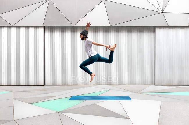 Ganzkörperbärtiger Typ in Sportkleidung schwingt die Arme und springt beim Yoga-Workout im geräumigen Raum mit geometrischen Ornamenten über den Boden — Stockfoto