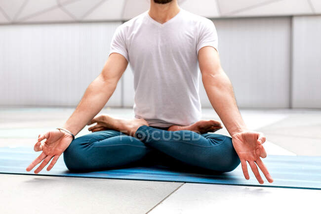 Обрезанный неузнаваемый мужчина в спортивной одежде, сидящий в позе Лотоса и медитирующий с закрытыми глазами и жестом мудры в просторной комнате с геометрической стеной и полом — стоковое фото