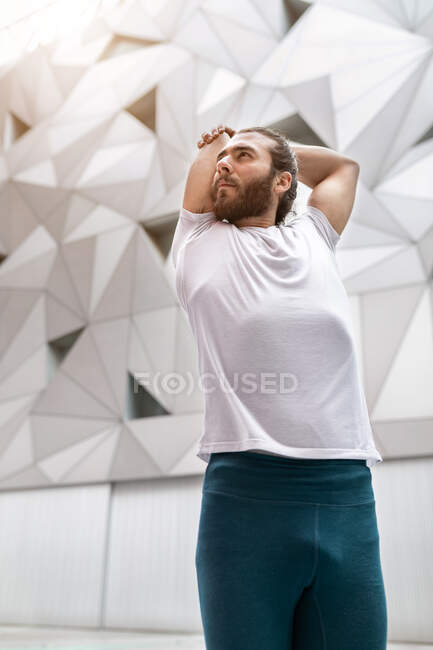 Desde abajo tipo barbudo en camiseta blanca mirando hacia otro lado y haciendo ejercicio de estiramiento para los brazos mientras hace entrenamiento de yoga contra la pared geométrica ornamental - foto de stock