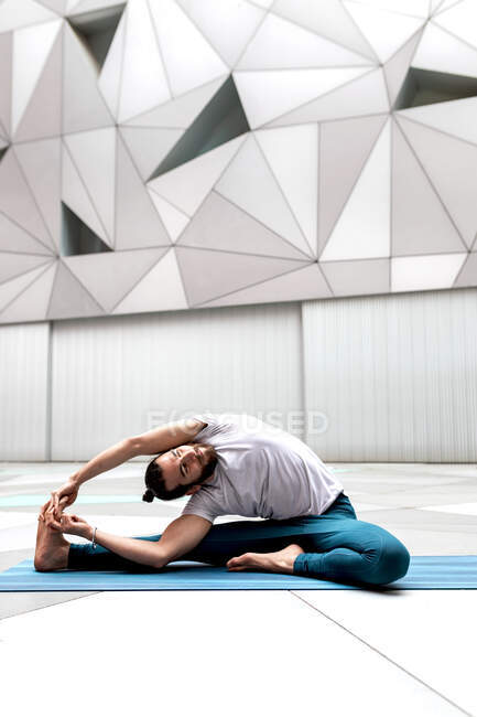Homme barbu pieds nus faisant Revolved Head to Knee exercice tout en étant assis sur le tapis contre le mur géométrique pendant l'entraînement de yoga — Photo de stock