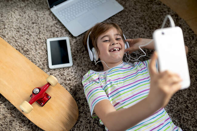 Зверху веселий маленький хлопчик у повсякденній сорочці слухає музику з навушниками, лежачи на килимі біля гаджетів і скейтборду в спальні — стокове фото