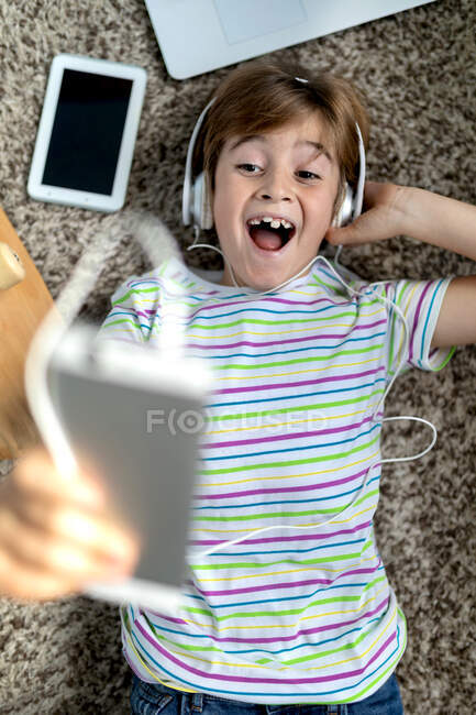 From above of cheerful little boy in casual shirt listening to music with headphones while lying on carpet near gadgets and skateboard in bedroom — Stock Photo