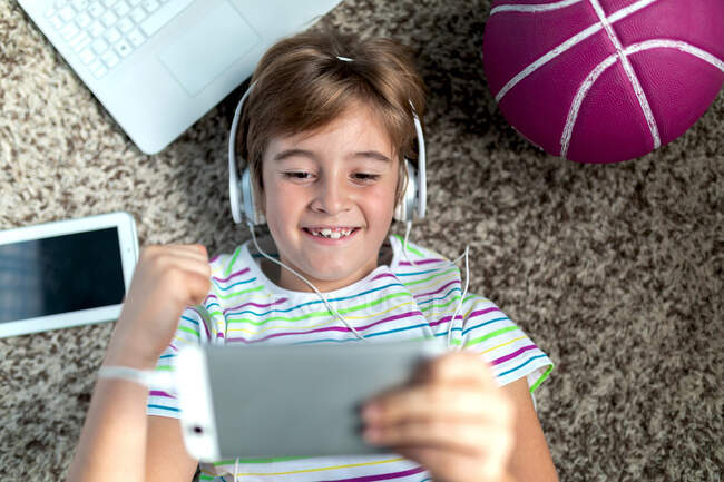 From above of cheerful little boy in headphones lying on floor near gadgets and ball and playing video game on tablet while spending time at home — Stock Photo