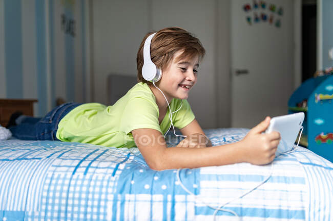 Позитивний хлопчик у повсякденному одязі з навушниками лежить на ліжку і дивиться фільм на планшеті, відпочиваючи вдома — стокове фото
