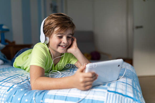 Positivo ragazzo preadolescente in abbigliamento casual con cuffie sdraiato sul letto e guardare film su tablet mentre si riposa a casa — Foto stock
