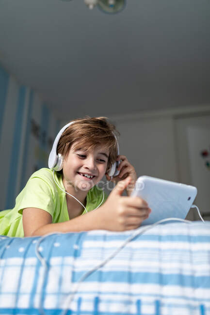 Positiver Frühchen-Junge in Freizeitkleidung mit Kopfhörern auf dem Bett liegend und Film auf Tablet anschauend, während er zu Hause ruht — Stockfoto