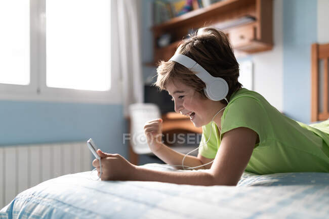 Menino pré-adolescente positivo em desgaste casual com fones de ouvido deitados na cama e assistindo filme em tablet enquanto descansa em casa — Fotografia de Stock