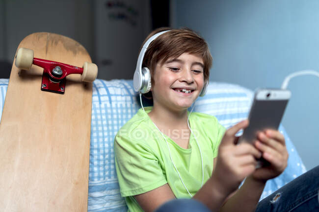 Ragazzino ridente con le cuffie che ascolta musica e chatta con gli amici sui social network mentre siede vicino allo skateboard in camera da letto — Foto stock