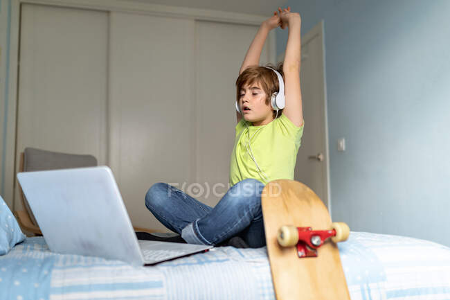 Niño deprimido con auriculares en el cuello sentado en la cama y el uso de la computadora portátil durante el aislamiento debido a coronavirus en casa - foto de stock