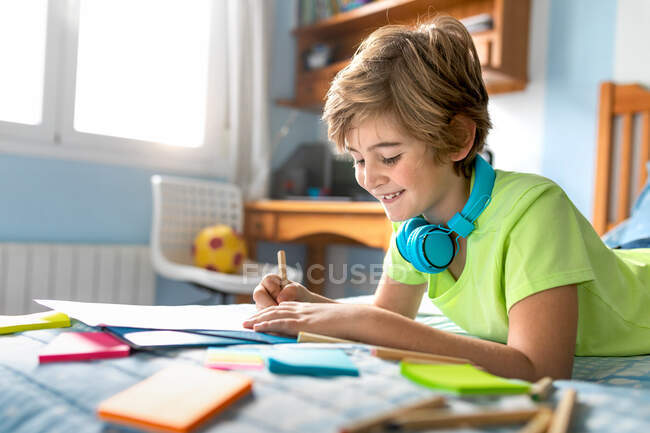 Side view of positive schoolboy in casual wear and wireless headphones drawing with pencils while spending free time in bedroom — Stock Photo