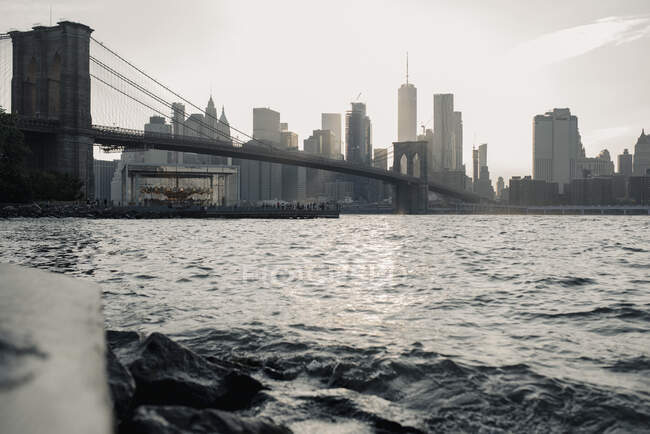 Удивительный низкоугловой городской пейзаж Нью-Йорка с Бруклинским мостом через реку и небоскребами под солнечным светом на закате — стоковое фото