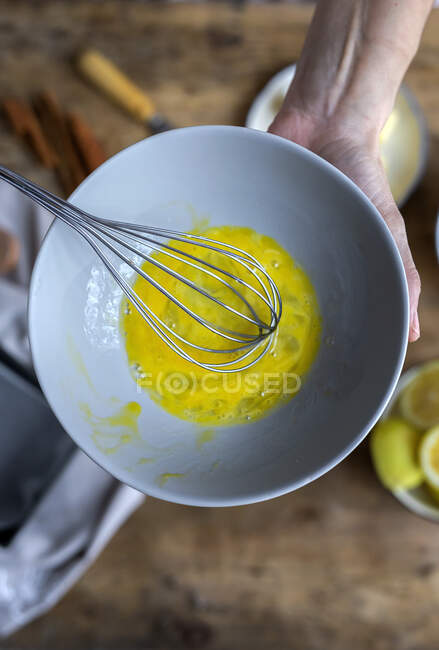 Сверху вид на урожай анонимная женщина взбивает яйца в черной миске на деревянном столе с лимоном, мукой, маслом и корицей палочки ингредиенты для торта — стоковое фото