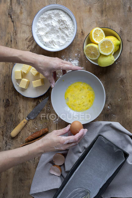Зверху невизначена жінка розбиває свіже куряче яйце в миску, готуючи тістечко на дерев'яному столі зі свіжими інгредієнтами — стокове фото