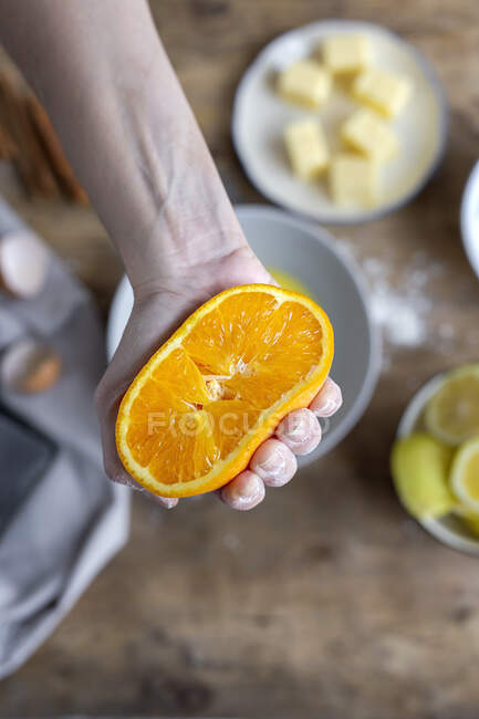 Зверху рука невизначеної жінки, вкрита борошном, тримає і показує камеру свіжий нарізаний на половину апельсин над мискою, готуючи тісто за столом — стокове фото