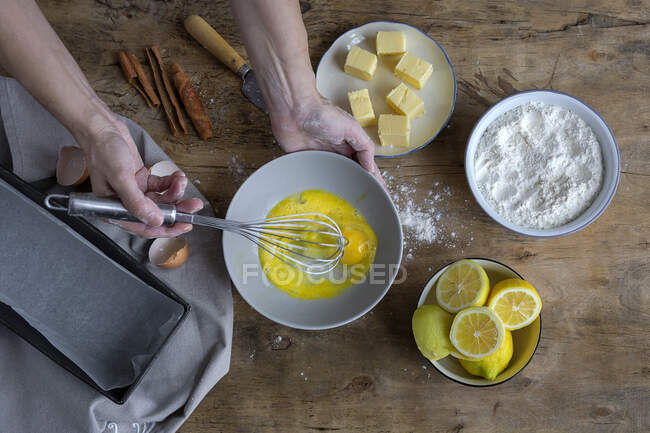 Vue du dessus de la récolte femme anonyme fouettant des œufs dans un bol noir sur une table en bois avec du citron, de la farine, du beurre et des bâtonnets de cannelle ingrédients pour gâteau — Photo de stock
