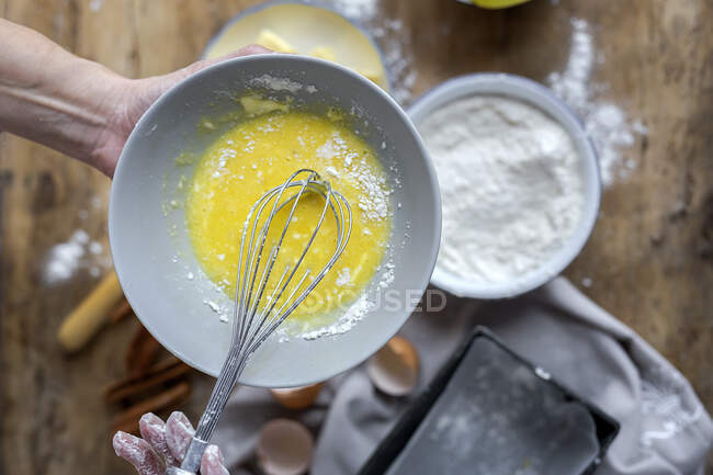 Desde arriba vista superior de la cosecha mujer anónima batiendo huevos en un tazón negro en una mesa de madera con limón, harina, mantequilla y palitos de canela ingredientes para la torta - foto de stock