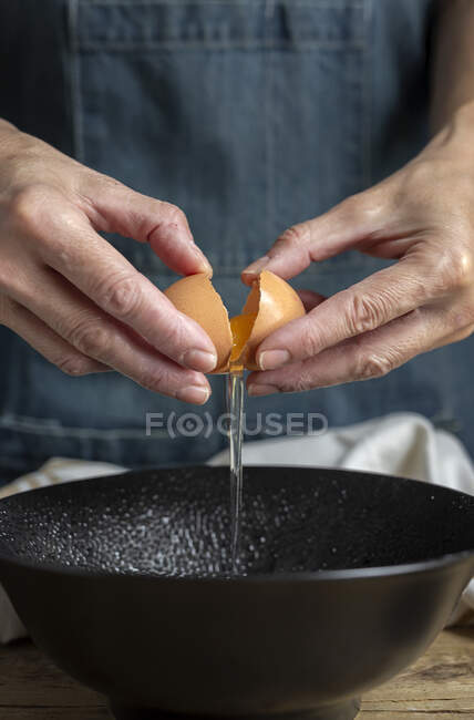 Hembra irreconocible rompiendo huevo de pollo fresco en un tazón mientras cocina pastelería en una mesa de madera con ingredientes frescos - foto de stock