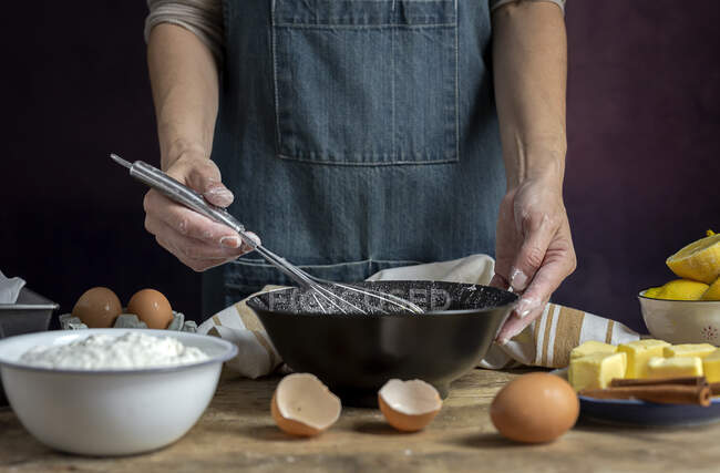 Coltiva anonima donna in grembiule montando uova in ciotola nera su tavolo di legno con limone, farina, burro e bastoncini di cannella ingredienti per la torta — Foto stock